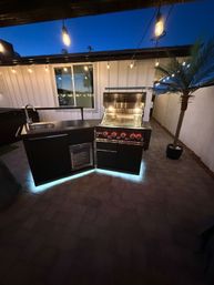 Backyard outdoor kitchen at dusk with illuminated stainless-steel gas grill, black prep island and sink on a paver patio, string lights overhead and a potted palm for a cozy tropical vibe.