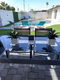 Sunny residential backyard pool patio with turquoise lounge chairs, glass dining table topped with two potted succulents and a QR-code sign, artificial turf and palm accents