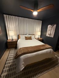 Cozy modern bedroom interior with white bedding and brown throw pillow, wooden nightstands and lamps, dark painted walls, ceiling fan, sheer curtains, horse wall art and patterned area rug