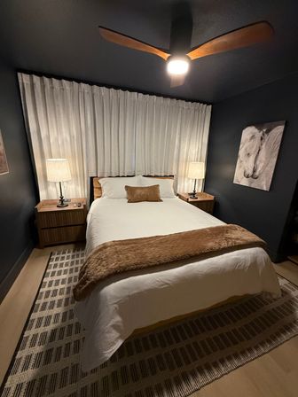 Cozy modern bedroom interior with white bedding and brown throw pillow, wooden nightstands and lamps, dark painted walls, ceiling fan, sheer curtains, horse wall art and patterned area rug