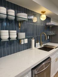 Sleek modern kitchen sink area with vertical navy tile backsplash, white quartz countertop, brass gooseneck faucet and sconce, glass floating shelves displaying stacked white dishes and clear glasses