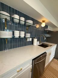 Modern kitchen countertop with navy vertical tile backsplash, glass floating shelves of white dishes, brass faucet over undermount sink, white quartz counter and stainless dishwasher