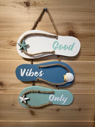 Hanging coastal wall decor showing three wooden flip-flop plaques on a natural wood wall reading “Good Vibes Only,” with rope straps, starfish and shell accents in teal and blue — beach house/nautical vibe.