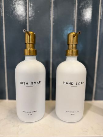 Chic minimalist white soap dispensers with brass pumps labeled "Dish Soap" and "Hand Soap" on a marble countertop in front of a dark blue subway tile backsplash.