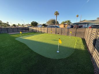 Sunlit suburban backyard putting green with artificial turf, five yellow flags, wooden slat fence, parked cars and palm trees under a clear blue sky