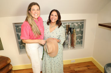 Two smiling women in a bright dressing nook — one in a pink striped shirt and white pants, the other in a mint patterned dress holding a tan hat, with scarves and accessories displayed on a recessed shelf behind them.