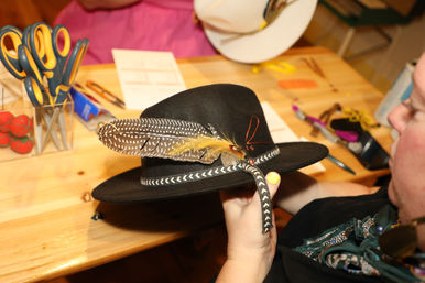 Black felt fedora with chevron braided band and spotted feather trim held above a wooden craft table with scissors and other hat-making supplies