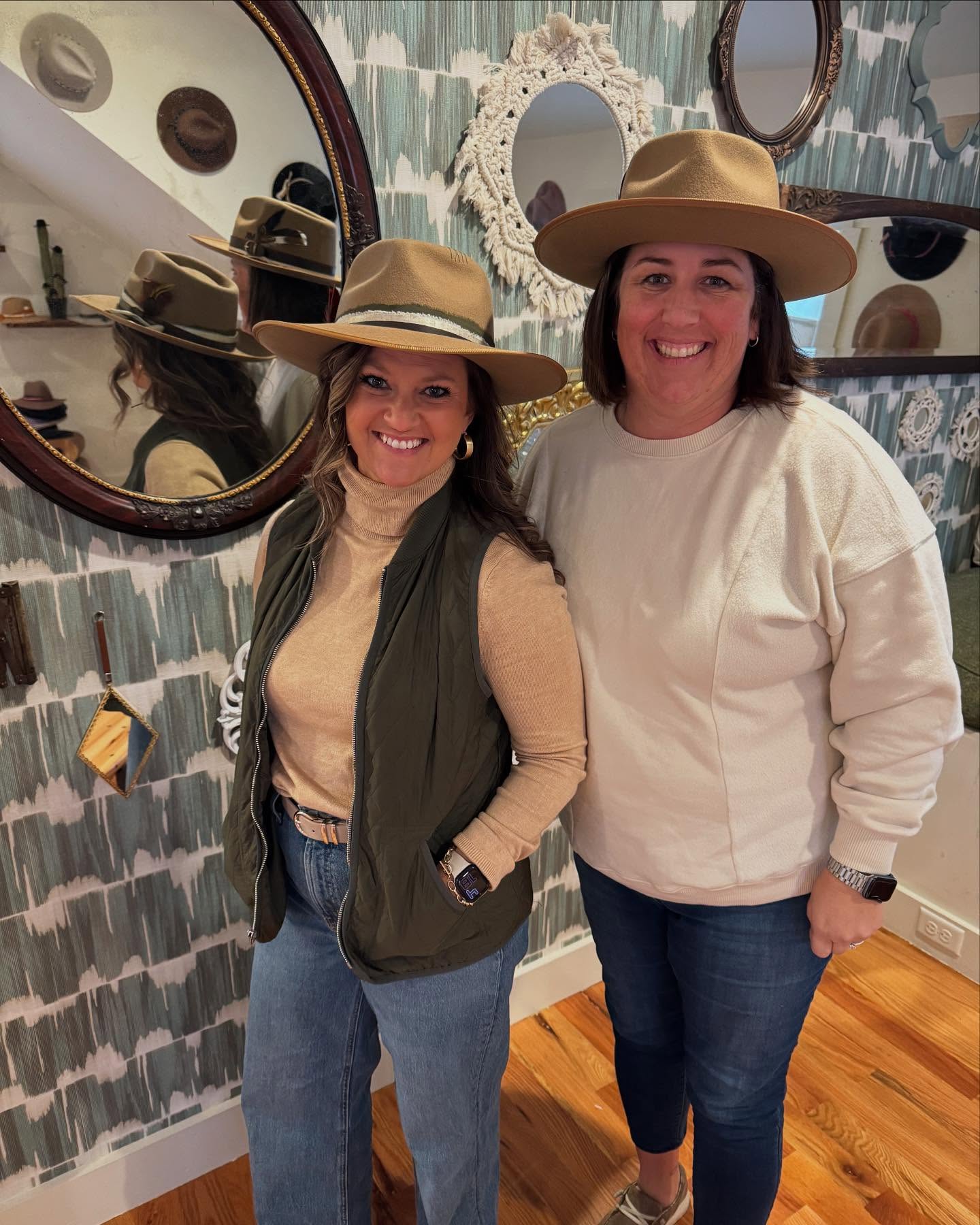 Two smiling women wearing tan wide-brim felt hats and casual fall outfits, posing inside a cozy hat shop with decorative mirrors, patterned wallpaper, and hardwood floors.