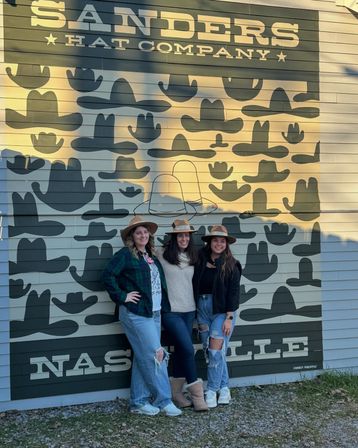 Three friends wearing wide-brim hats smile and pose in front of a large hat-pattern mural on an outdoor painted wall in Nashville