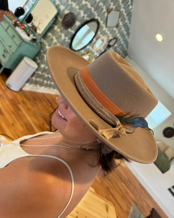 Side profile of a woman wearing a tan felt fedora with an orange ribbon and feather, smiling in a sunlit boho-style room with hardwood floors and decorative mirrors.