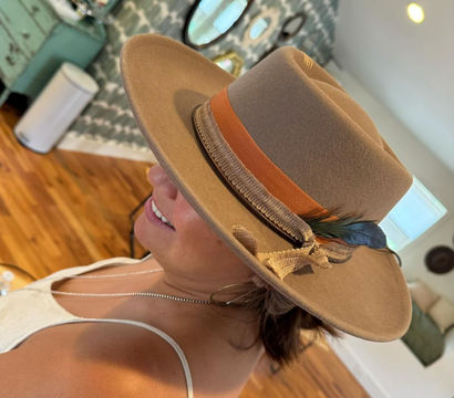 Side profile of a woman wearing a tan felt fedora with an orange ribbon and feather, smiling in a sunlit boho-style room with hardwood floors and decorative mirrors.