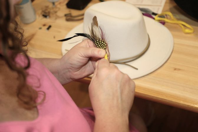Hands attaching decorative feathers and a beaded accent to a white wide-brim hat on a wooden craft table