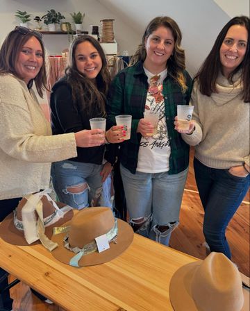 Four smiling women in a cozy boutique loft toasting with drinks over a wooden table showcasing tan felt hats with decorative ribbons and bands; casual outfits include knit sweaters, flannel shirt, and ripped jeans.