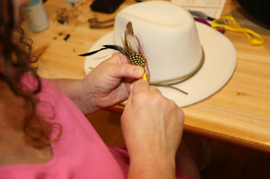 Close-up of hands crafting a DIY feather embellishment with spotted and long feathers beside a wide-brim white hat on a wooden table.