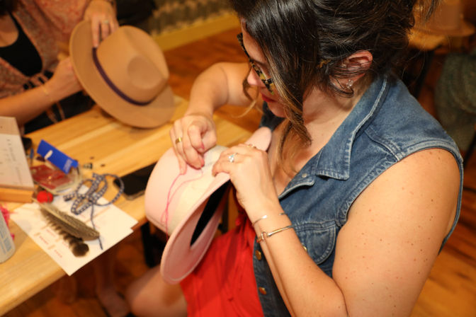 Close-up of a person hand-sewing and decorating a pink felt hat at a wooden craft table in a hat-making workshop, with a tan hat, ribbons, feathers, and tools nearby.