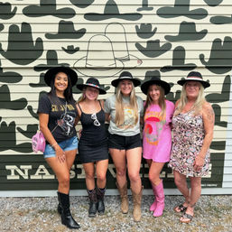 Five women wearing cowboy hats and boots smiling and posing in front of a hat-pattern mural on a Nashville street, casual summer outfits and country-style accessories.