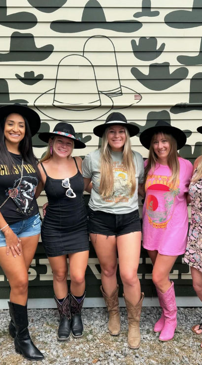 Five women wearing cowboy hats and boots smiling and posing in front of a hat-pattern mural on a Nashville street, casual summer outfits and country-style accessories.