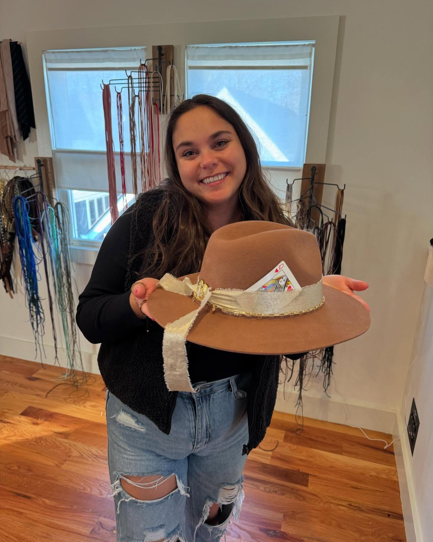 Smiling woman in ripped jeans holding a brown felt wide-brim hat with a cream ribbon and tucked playing card, standing in an accessory boutique with hanging leather cords and hardwood floors