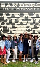 Seven friends wearing cowboy-style hats and casual outfits pose, smiling in front of a large Western hat-patterned mural on an outdoor storefront wall.