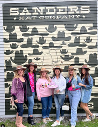 Six friends wearing wide-brim hats smiling and posing in front of a large hat-themed storefront mural covered in cowboy hat silhouettes, holding drinks for a fun outdoor group photo.