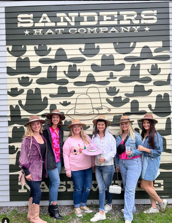 Six friends wearing wide-brim hats smiling and posing in front of a large hat-themed storefront mural covered in cowboy hat silhouettes, holding drinks for a fun outdoor group photo.