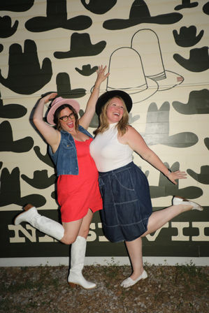 Two women in hats and summer dresses strike playful kicked-leg poses in front of a large black cowboy-hat mural on an outdoor wall.