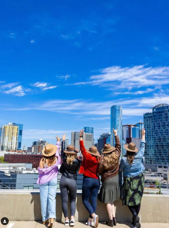 Five friends in casual outfits and hats stand on a rooftop with arms raised, overlooking a downtown city skyline under a bright blue sky with wispy clouds.