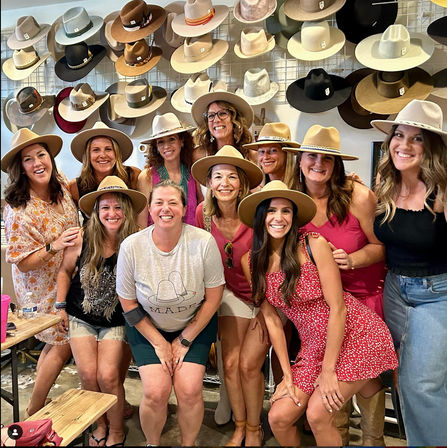 Smiling group of women trying on wide‑brim and cowboy-style hats, posing inside a hat boutique in front of a wall display of neutral-toned fedoras and Western hats.