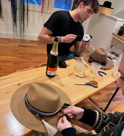 Hands attaching a braided band and tassel to a brown felt fedora in a DIY hat-making workshop on a wooden table with scissors, ribbon spool, pliers, a champagne bottle and plastic cups in a craft studio.