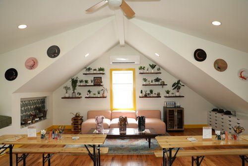 Cozy sunlit attic loft lounge with vaulted white ceiling and fan, pink sofa beneath a bright window, wall shelves of potted plants, hats displayed on sloped walls, long wooden communal tables and hardwood floors