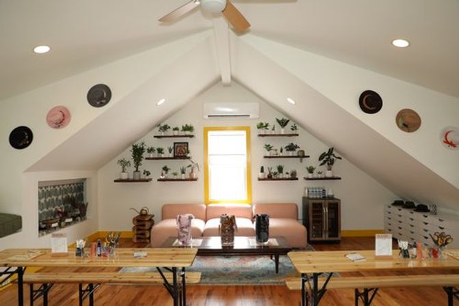 Cozy sunlit attic loft lounge with vaulted white ceiling and fan, pink sofa beneath a bright window, wall shelves of potted plants, hats displayed on sloped walls, long wooden communal tables and hardwood floors