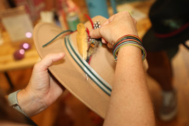 Tan felt fedora with green-striped ribbon and feather embellishment being adjusted by hands wearing colorful stacked bracelets in a shop setting