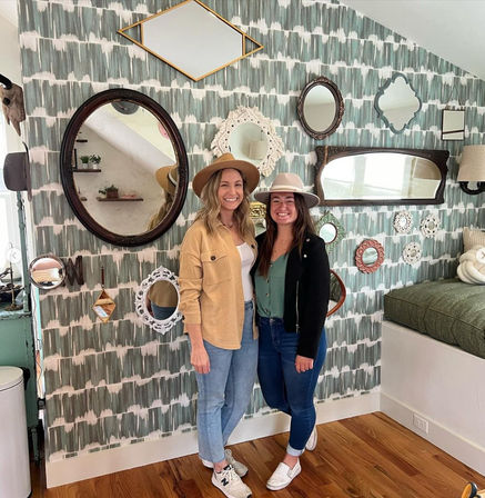 Two smiling women wearing hats pose in front of a gallery wall of assorted vintage mirrors on green patterned wallpaper in a cozy home-decor setting.