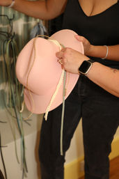 Close-up of a person holding a playful pink felt cowgirl hat with braided gold trim, pale ribbon and dangling cords in a boutique accessory display.