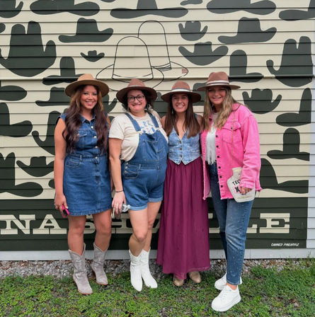 Four people in cowboy hats and western-style outfits — denim dress, overalls, long skirt and pink jacket — smiling and posing in front of a painted cowboy-hat mural on building siding.