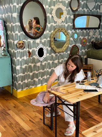 Person seated at a wooden table in a colorful boutique studio adjusting a lavender wide‑brim hat; patterned wallpaper with a gallery of decorative mirrors, hardwood floor and scattered craft supplies.
