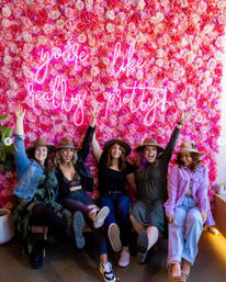 Five friends in hats cheer and kick their feet in front of a vibrant pink rose wall with neon script reading “you’re like really pretty” — an Instagrammable floral photo backdrop.