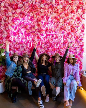 Five friends in hats cheer and kick their feet in front of a vibrant pink rose wall with neon script reading “you’re like really pretty” — an Instagrammable floral photo backdrop.