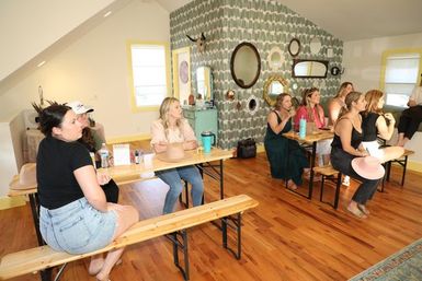 Sunlit loft studio workshop with a group of women seated on wooden benches at tables, holding hats and drinks, facing a patterned gallery wall of mirrors and yellow‑trimmed windows on hardwood floors.