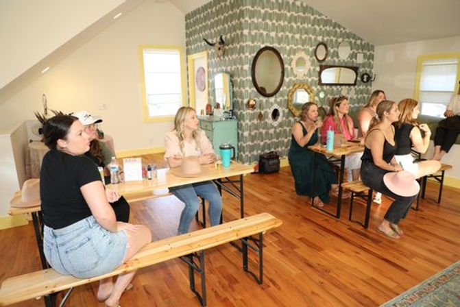 Sunlit loft studio workshop with a group of women seated on wooden benches at tables, holding hats and drinks, facing a patterned gallery wall of mirrors and yellow‑trimmed windows on hardwood floors.