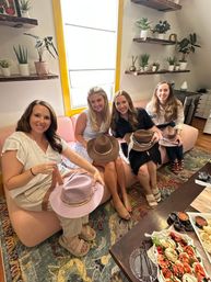 Four women on a pink sofa in a bright, plant-filled studio trying on hats during an indoor brunch with charcuterie and caprese salad on the coffee table.
