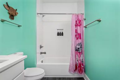 Cheerful turquoise bathroom with white tub and shower, pink patterned shower curtain, chrome fixtures, white vanity and toilet, dark tile floor, whimsical wooden animal head on the wall.