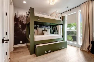 Sunlit modern bedroom with olive-green bunk bed and pull-out trundle, forest mural accent wall, hardwood floors and large floor-to-ceiling window.