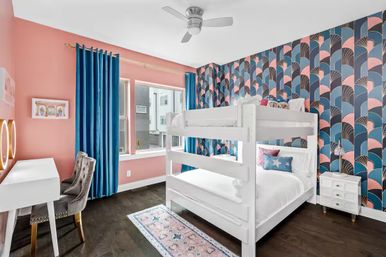 Stylish kids' bedroom in an apartment with a white bunk bed against blue-and-pink art-deco wallpaper, coral-pink walls, deep-blue velvet curtains, dark hardwood floors, white desk with tufted gray chair, pink patterned rug and small white nightstand.