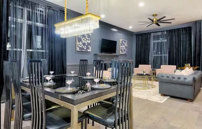 Glam open-plan interior: glass-top dining table set for six with tall black chairs, gold crystal chandelier, floor-to-ceiling dark curtains, gray tufted sofa and blush accent chairs on light wood floors.