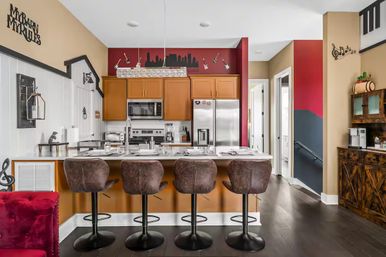 Open-concept modern kitchen with breakfast bar and four brown swivel stools, white island set with place settings, stainless steel appliances and warm wood cabinets, red accent wall with city-skyline decal and guitar decor, dark hardwood floors and music-themed accents.
