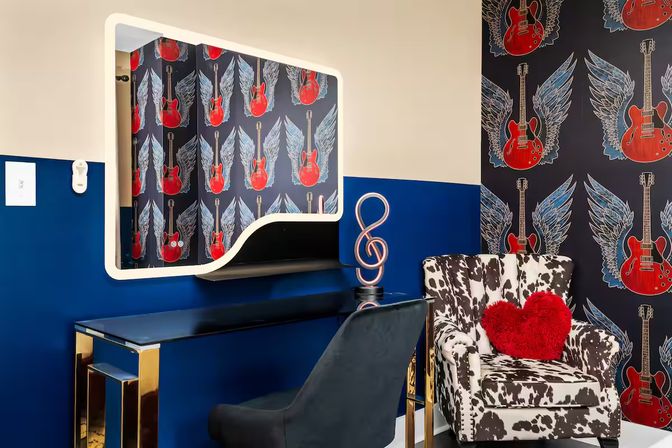 Music-themed room corner with navy half-wall and winged red guitar wallpaper, white-framed mirror above glossy desk with gold legs, black velvet chair, cow-print armchair with red heart pillow and treble-clef lamp.