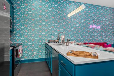 Bright tropical-inspired kitchen with teal cabinets, white quartz island and sink, pink-and-teal bird-patterned wallpaper, neon “Be Flamingo” sign and a casual breakfast bar with place settings.