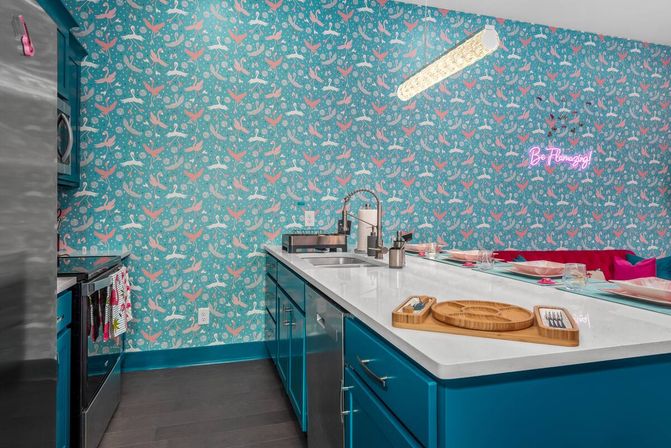 Bright tropical-inspired kitchen with teal cabinets, white quartz island and sink, pink-and-teal bird-patterned wallpaper, neon “Be Flamingo” sign and a casual breakfast bar with place settings.