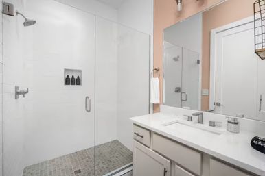 Bright modern bathroom with frameless glass shower, rain showerhead and recessed niche with black bottles, gray tiled shower floor, white vanity with quartz countertop, rectangular mirror and peach accent wall.